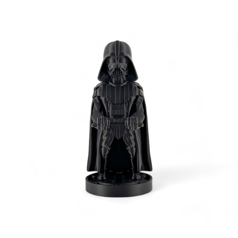 May include: A black plastic figurine of Darth Vader from Star Wars. The figurine is standing with its arms crossed and is wearing a black cape. The figurine is on a black base.