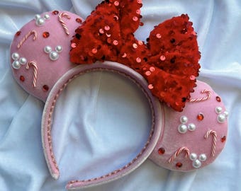 May include: Pink mouse ear headband with a red sequined bow. The ears are decorated with candy canes and faux pearls. The headband is trimmed with pink fabric.