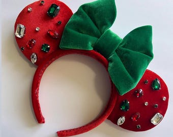 May include: Red velvet mouse ear headband with a large green bow. The ears are adorned with various colorful gemstones. The headband is a festive accessory, perfect for themed events or everyday wear.