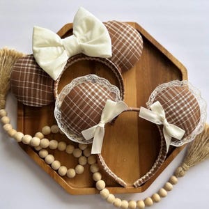 May include: Two headbands with round ears featuring a brown and white plaid fabric pattern. One headband has a large cream-colored fabric bow. The other headband features white lace trim around the ears and two small cream-colored ribbon bows.