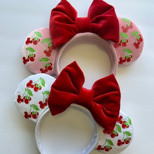 May include: A pair of white and pink Minnie Mouse ears with red velvet bows. The ears are decorated with a cherry print.