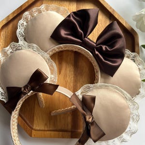 May include: Several headbands with beige padded ears, adorned with brown satin bows and delicate lace trim. The headbands are arranged on a wooden tray, showcasing their elegant design and craftsmanship.