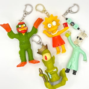 May include: Four character keychains featuring a green creature with orange flippers, a yellow girl with a red dress, a green one-eyed alien with a crown, and a light green scientist figure with black glasses and a beaded chain attachment.