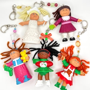 May include: Five Cabbage Patch Kids doll keychains with yarn hair and colorful outfits. One wears a white dress, one a purple dress, one a pink and blue dress with a yellow bear, one a green skating outfit, and one a red and green apron outfit.