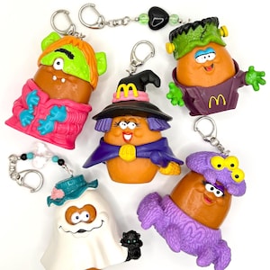 May include: Five McDonald's Halloween-themed potato character keychains. One wears a green mask and pink jacket, one a witch hat and purple cape, one a Frankenstein costume, one a white ghost sheet with a blue hat, and one a purple monster suit.