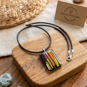 Peut inclure: Un collier avec un pendentif rectangulaire et un cordon noir. Le pendentif transparent présente des lignes verticales colorées jaunes, oranges, rouges et blanches. Le collier est présenté sur une surface en bois. Une carte indique "SACS & PATOUNES".