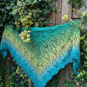 Może przedstawiać: Ręcznie robiona chusta w odcieniach turkusu, zieleni i żółci, zawieszona na płocie. Chusta ma delikatny, ażurowy wzór koronki. Przejście kolorów od żółtego do zielonego do turkusowego tworzy atrakcyjny wizualnie dodatek.