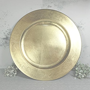 Könnte beinhalten: Ein goldfarbener Platzteller mit Metallic-Finish. Der Teller hat einen breiten Rand und eine leicht vertiefte Mitte. Die Oberfläche hat eine gebürstete Textur, die ihm einen dezenten Glanz verleiht. Der Teller ist für dekorative Zwecke bestimmt.
