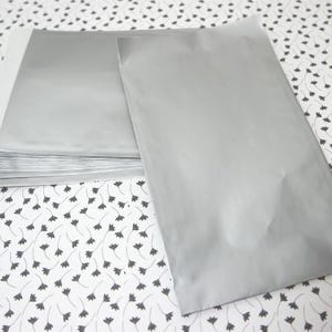 Könnte beinhalten: Ein Stapel silberfarbener, rechteckiger Verpackungsumschläge. Ein Umschlag ist separat, ein weiterer teilweise sichtbar. Die Umschläge liegen auf einer weißen Oberfläche mit einem schwarzen Blumenmuster.