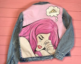 May include: A blue denim jacket with a pink painted design on the back. The design features a woman with pink hair crying and a thought bubble above her head with a slice of pizza inside. The text "@sonyashnick_jeans" is at the bottom of the design.