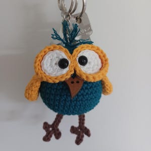 May include: A handmade crochet owl keychain. The owl has a teal body, white eyes with black pupils, and orange and yellow accents. The keychain is attached to a silver metal ring.