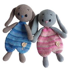 May include: Two handmade crochet bunny dolls. One bunny is gray with blue overalls, the other is gray with pink overalls. Both have button details and are holding hands. The bunnies have long ears and closed eyes.