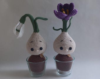 Può includere: Due figure di piante all'uncinetto fatte a mano. Una ha un fiore bianco, l'altra un fiore viola. Entrambe hanno corpi beige a forma di bulbo con occhi a bottone e sono poste in piccoli bicchieri di vetro. Le piante sono su sfondo bianco.