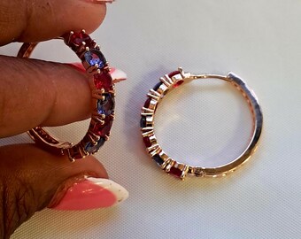 May include: A pair of hoop earrings crafted from rose-toned metal, each featuring an alternating pattern of round-cut red and blue gemstones set along the outer edge. The earrings have a hinged closure mechanism and a polished metallic finish.