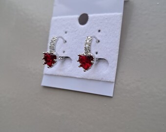 May include: A pair of silver-toned hoop earrings with a heart-shaped, deep red gemstone. The hoops are embellished with small, clear crystals. The earrings are displayed on a white card.