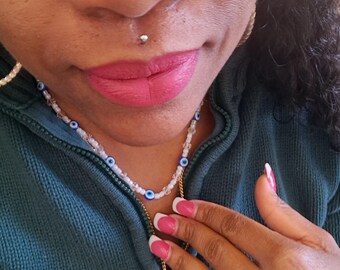 May include: A close-up of a person wearing a white beaded necklace with blue evil eye beads and a gold chain necklace with a gold Hamsa hand pendant. The person is wearing a teal green zip-up jacket and has pink lipstick and pink and white French tip nails.