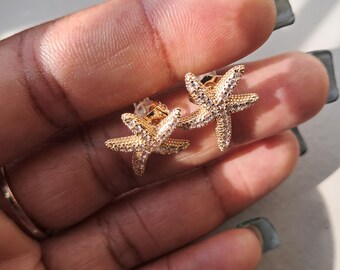 May include: A pair of gold-colored starfish stud earrings. Each earring features a starfish design encrusted with small, clear crystals. The earrings are displayed against a blurred background.