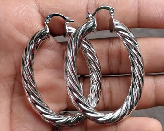 May include: A pair of silver-colored hoop earrings with a twisted rope design. The earrings are oval-shaped and have a shiny, metallic finish. The hoops are held in a hand, showcasing their size and detail.