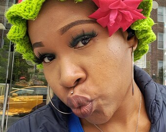 May include: A green, white, and red crocheted hat with a ruffled brim and a pink flower accent. The hat is worn with a silver necklace with a heart-shaped pendant and hoop earrings. The person is wearing a blue top and a black jacket.