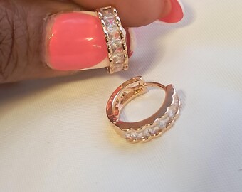 May include: A pair of rose gold hoop earrings featuring a row of clear, square-cut stones embedded along the front surface. The hoops have a hinged closure mechanism and a scalloped edge design along the sides, showcasing a metallic finish.