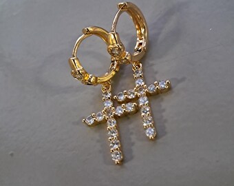 May include: A pair of gold-colored hoop earrings with cross pendants. Each cross is encrusted with small, clear gemstones. The earrings are displayed against a neutral gray background.