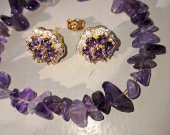 May include: A jewelry set featuring a gold ring and earrings with purple and white gemstones, along with a necklace made of purple amethyst stones. The earrings have a flower design. The jewelry is set against a neutral background.