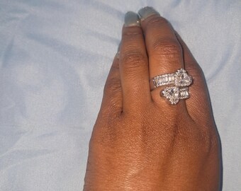 May include: A silver-toned bypass ring featuring two heart-shaped center stones surrounded by small pavé-set crystals. The band is split into two sections, each adorned with a row of baguette-cut crystals, creating a layered and sparkling appearance.