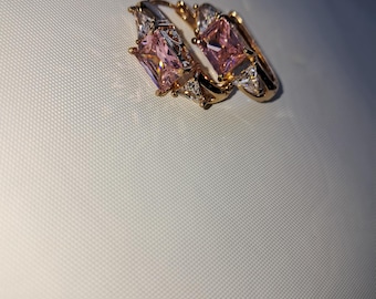 May include: A pair of gold-toned hoop earrings featuring a central rectangular pink gemstone flanked by two smaller triangular clear stones on each side. The metal setting is polished and reflective, showcasing a classic hinged closure design.