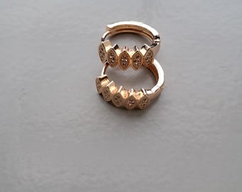 May include: A pair of rose gold hoop earrings. Each earring features a series of diamond-shaped embellishments with small, clear stones. The earrings are displayed on a light gray surface.