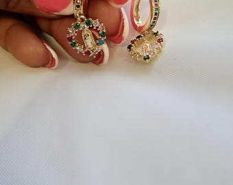 May include: A pair of gold-tone hoop earrings featuring a dangling heart-shaped charm. Each charm contains a central figure of the Virgin Mary surrounded by small, multi-colored gemstones. The hoops are also accented with a row of colorful stones.