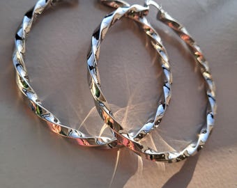 May include: A pair of silver-colored hoop earrings with a twisted rope design. The earrings are oval-shaped and reflect light, creating highlights and shadows. The earrings are displayed on a neutral-colored surface.