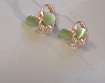 May include: A pair of gold-colored hoop earrings. Each earring features a light green, oval-shaped stone and a cluster of small, clear crystals. The earrings are designed with a floral or leaf-like motif.