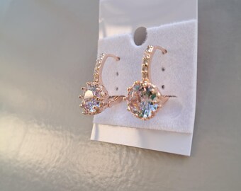 May include: Rose gold-colored hoop earrings with a large, clear, round gemstone as the focal point. The hoops are encrusted with smaller, clear gemstones. The earrings are displayed on a white card.