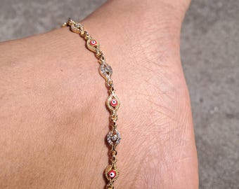 May include: Gold-toned anklet featuring alternating red and white eye-shaped charms. The anklet is adorned with clear, sparkling accents and is worn on an ankle. The jewelry is displayed outdoors in natural light.