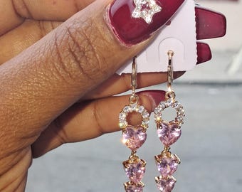 May include: Gold-toned dangle earrings with pink heart-shaped gemstones. The earrings feature a circular top with clear stones and a series of heart-shaped gems that cascade down. The earrings are displayed on a white earring card.