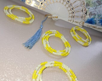 May include: Several strands of yellow and white beaded waist beads are displayed, along with a decorative white fan with gold accents and a blue tassel. The beads are arranged in circular patterns.
