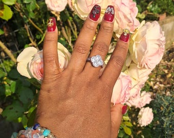 May include: A hand adorned with a flower-shaped ring and multiple bracelets. The nails are painted a deep red with decorative accents, including a gold cross, a snake, and a bow. The background features soft pink roses.