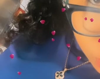 May include: A gold-colored necklace with a clover-shaped pendant, set with black stones, hangs against a blue top. The image includes black hair, glasses, and floating pink flower petals.