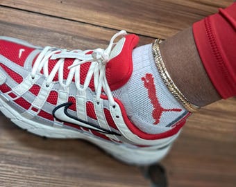 May include: Red, white, and gray athletic shoe with white laces and a black Nike swoosh. The shoe is paired with white socks featuring a red logo and a gold chain bracelet. The shoe rests on a wooden surface.