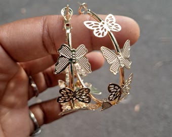 May include: Gold-colored hoop earrings featuring a butterfly design. The earrings have a series of butterfly cutouts around the hoop. The earrings are held up by a person's hand.