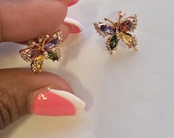 May include: A pair of butterfly-shaped stud earrings crafted from a rose gold-toned metal. Each earring features four marquise-cut multicolored gemstones in shades of purple, red, yellow, and green, accented with small clear stones along the wings.