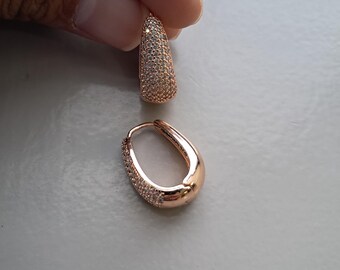 May include: Rose gold oval hoop earrings, one held by a hand. The top portion of one earring is covered in small, clear stones. The other earring is open, with a similar stone pattern along the outer edge.