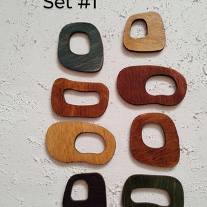 Op de afbeelding: Set van acht houten vormen met uitgesneden centra, geschilderd in verschillende kleuren, waaronder blauw, groen, bruin en zwart. De vormen zijn in twee rijen gerangschikt op een witte, getextureerde achtergrond. De tekst "Set #1" staat boven de vormen.