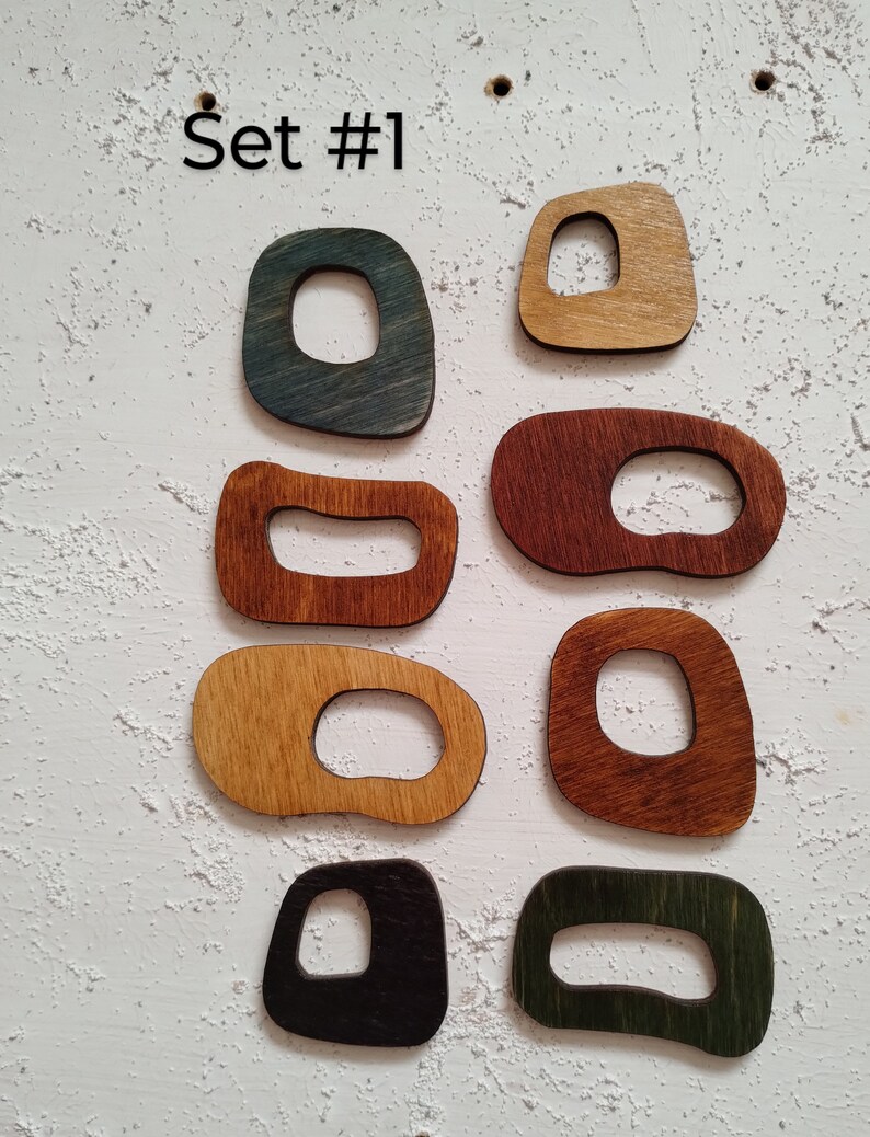 Op de afbeelding: Set van acht houten vormen met uitgesneden centra, geschilderd in verschillende kleuren, waaronder blauw, groen, bruin en zwart. De vormen zijn in twee rijen gerangschikt op een witte, getextureerde achtergrond. De tekst "Set #1" staat boven de vormen.