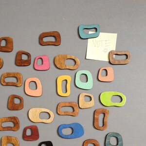 Op de afbeelding: Een verzameling kleurrijke houten vormen, waaronder cirkels, ovalen en rechthoeken, zijn gerangschikt op een grijs oppervlak. De vormen zijn in verschillende kleuren, waaronder blauw, groen, geel, roze en bruin. Een plakbriefje met het woord "NOTE" erop is bevestigd aan het oppervlak.