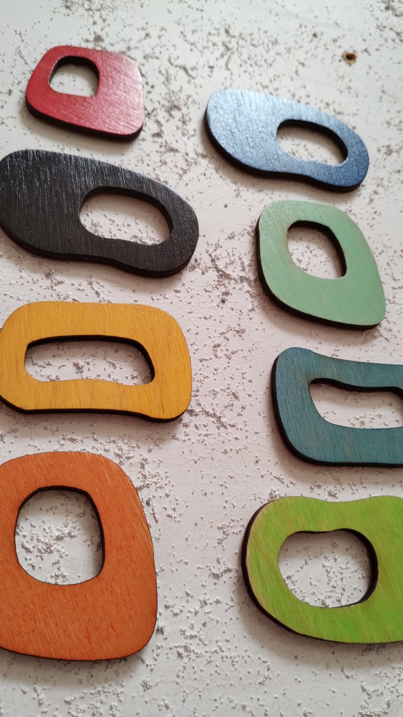 Op de afbeelding: Zeven houten vormen in verschillende kleuren met een gat in het midden. De vormen zijn rood, zwart, groen, geel, blauw, oranje en groen.
