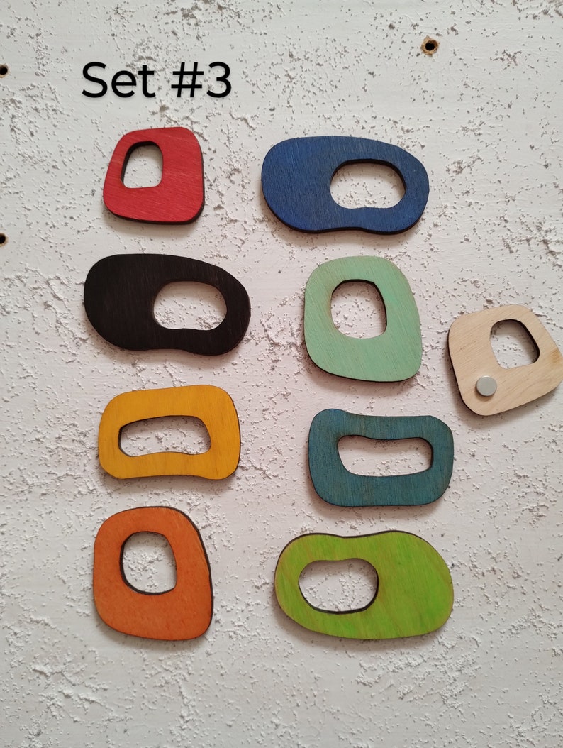 Op de afbeelding: Set van tien uitgesneden houten vormen in verschillende kleuren en ontwerpen. De vormen zijn allemaal ongeveer even groot en hebben een gat in het midden. De kleuren omvatten rood, blauw, zwart, groen, geel, oranje en bruin.