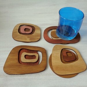 Op de afbeelding: Set van vier houten onderzetters met een geometrisch ontwerp. De onderzetters zijn in tinten bruin en roze. Een blauw glas staat op een van de onderzetters.