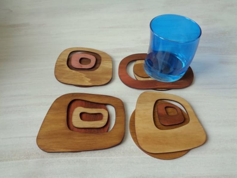 Op de afbeelding: Set van vier houten onderzetters met een geometrisch ontwerp. De onderzetters zijn in tinten bruin en roze. Een blauw glas staat op een van de onderzetters.
