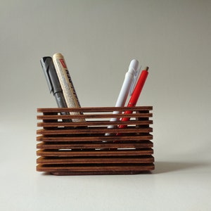 Puede incluir: Un portaplumas de madera con un diseño apilado. El portaplumas está hecho de madera marrón oscura y tiene una forma rectangular. Hay cuatro plumas dentro del portaplumas, incluyendo una pluma negra, una pluma gris, una pluma blanca y una pluma roja.